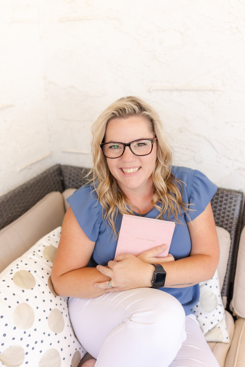 A person with long blonde hair and glasses is sitting on a cozy patio sofa, holding a pink notebook. They are wearing a blue top and white pants, surrounded by polka dot cushions. The background is a light, textured wall.