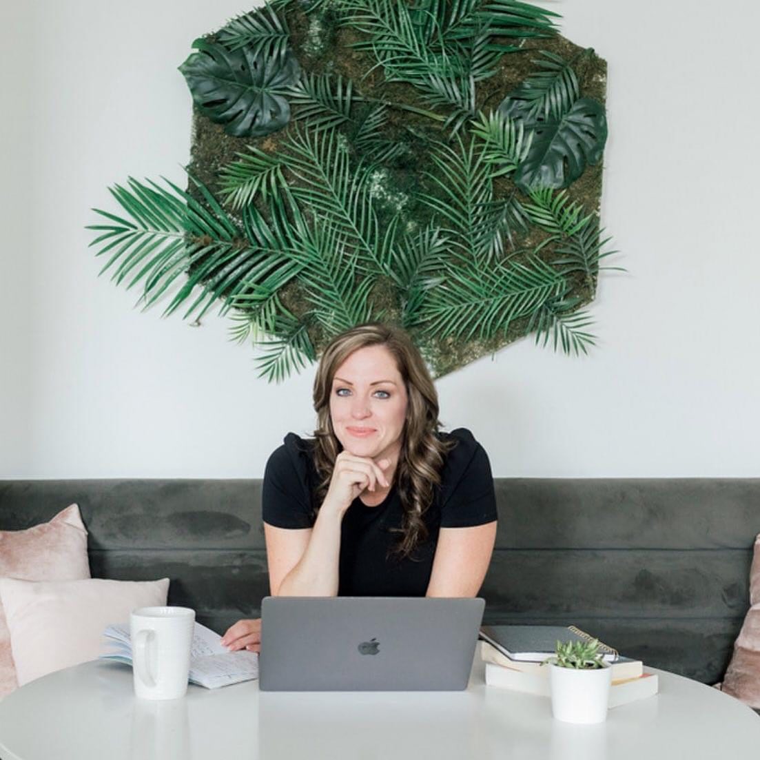 A woman sits at a table with a laptop, coffee mug, and books. Behind her is a decorative wall display of green leaves. She rests her chin on her hand and smiles as she promotes black friday deals for photographers 