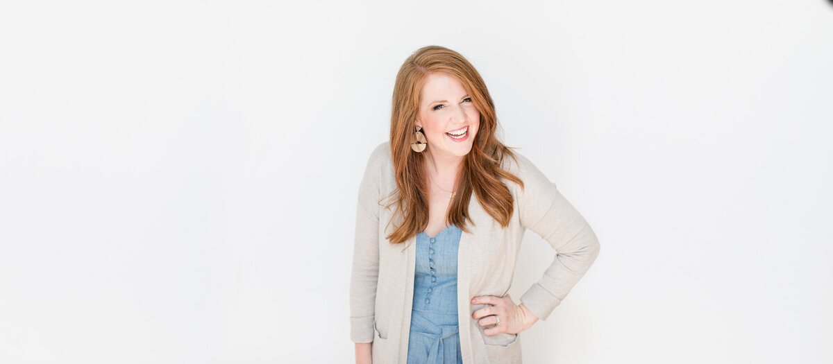 Woman with long red hair smiling, wearing a light gray cardigan over a blue dress, stands against a plain white background. She is looking at the camera with her hand on her hip while promoting black friday deals for photographers .