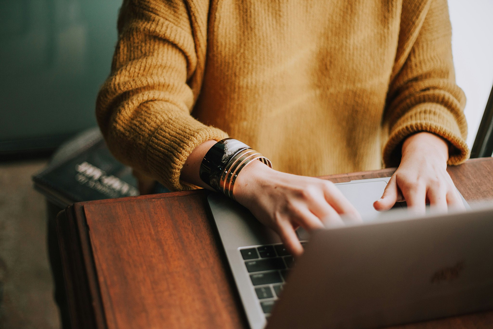 A woman sits at a wooden table typing in a mac laptop in a yellow sweater