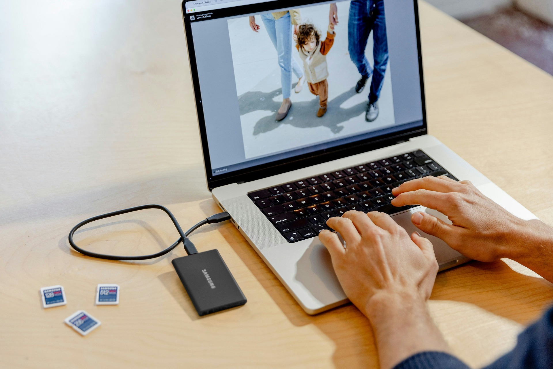 A look over the shoulder of someone editing photos on a laptop with a hardrive and sd cards before discovering Aftershoot vs Imagen