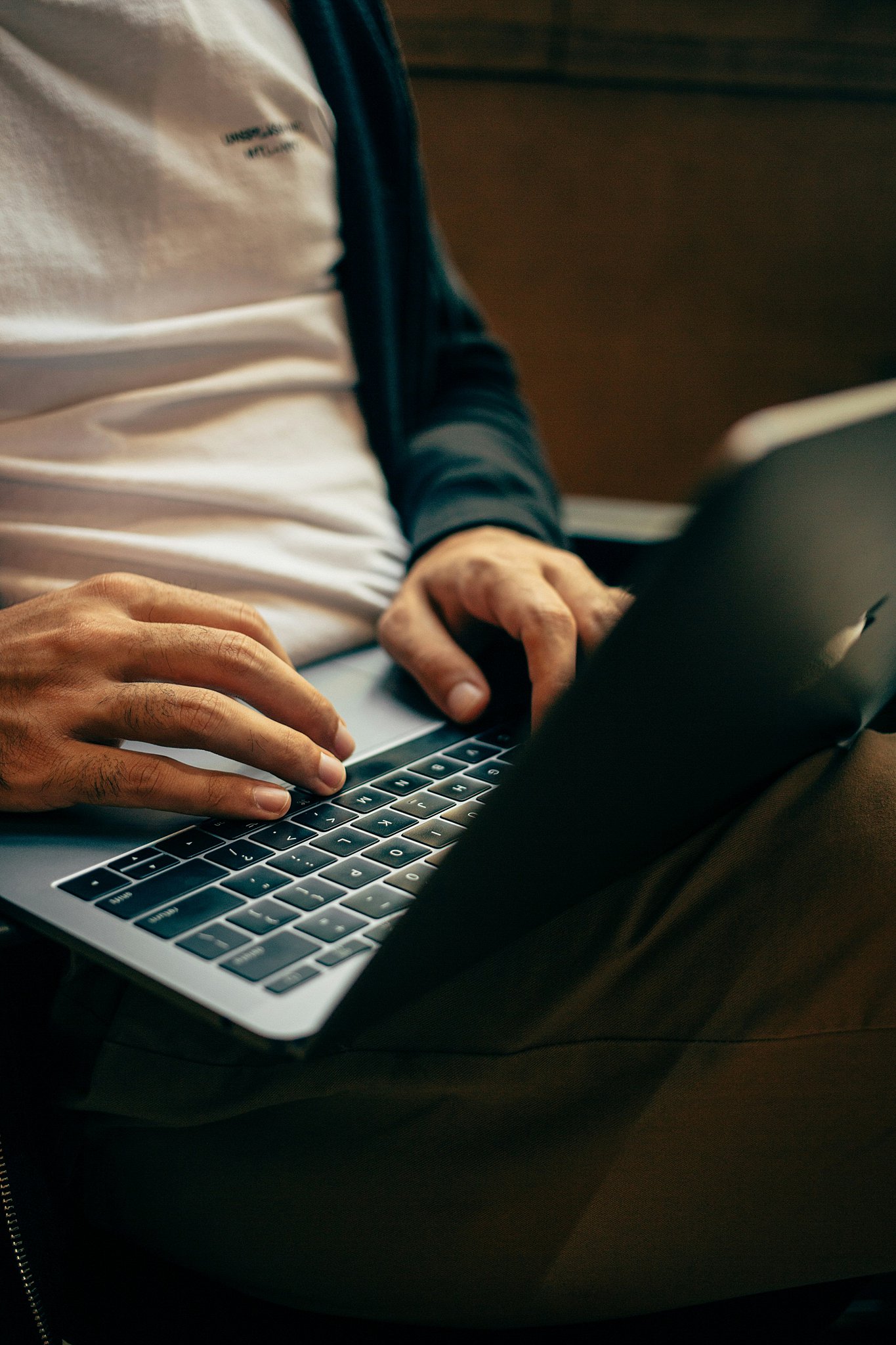 Details of a man typing on a mac laptop in his lap in a hoodie while researching Aftershoot vs Imagen
