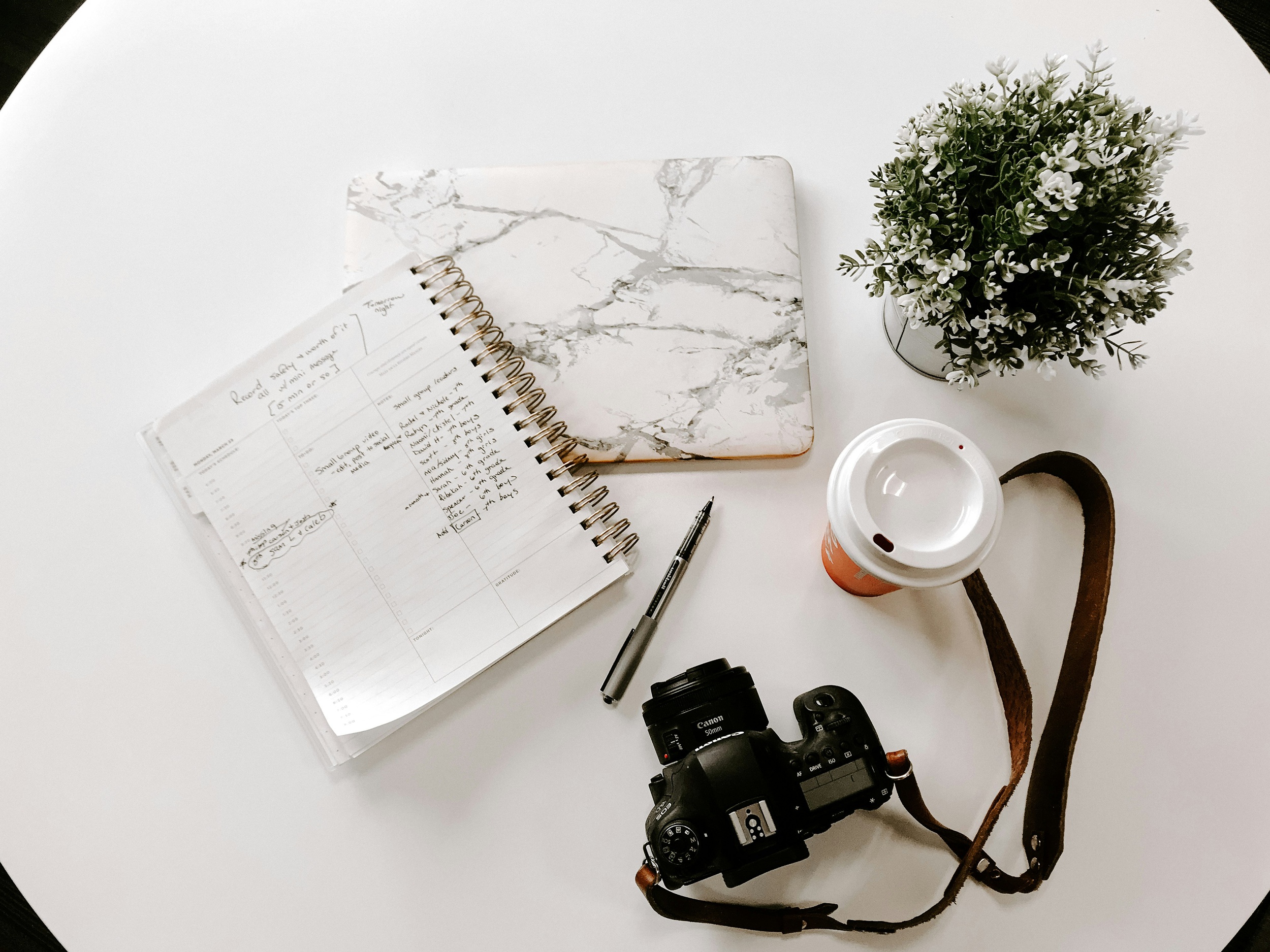 Details of a camera, coffee, laptop, calendar and plant on a white desk as a photographer looks into outsource blog writing